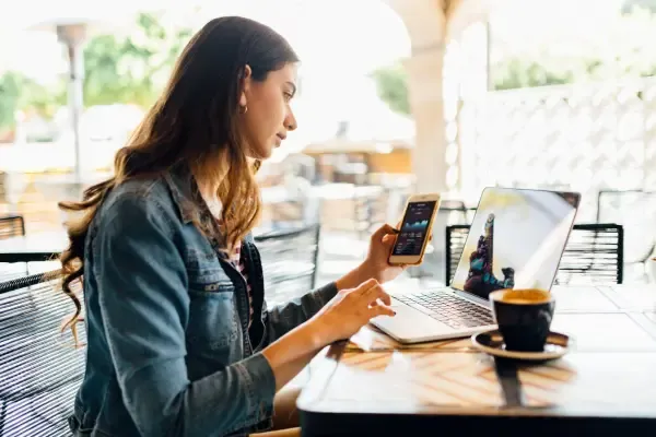 A woman sits at a cafe table, checking her phone while working on a laptop with a cup of coffee nearby